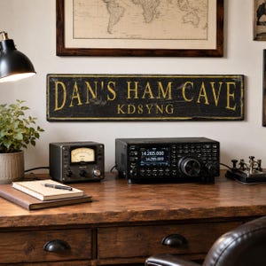 May include: A rectangular black sign with distressed edges and yellow lettering that reads DAN'S HAM CAVE and KD8YNG. The sign is mounted on a wall above a wooden desk featuring a radio transceiver, a signal meter, and a spiral-bound notebook.