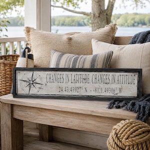 May include: A rectangular wooden sign with a distressed white finish and a black frame. The sign features a black compass rose graphic on the left and the text "CHANGES IN LATITUDE, CHANGES IN ATTITUDE" followed by the coordinates "24.43.43927 N - 81.1.49306 W".