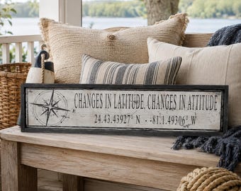 May include: A rectangular wooden sign with a distressed white finish and a black frame. The sign features a black compass rose graphic on the left and the text "CHANGES IN LATITUDE, CHANGES IN ATTITUDE" followed by the coordinates "24.43.43927 N - 81.1.49306 W".