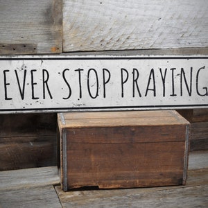 Puede incluir: Un letrero de madera blanco con texto negro que dice "NEVER STOP PRAYING." Una silueta negra de manos en oración está en el lado derecho del letrero.