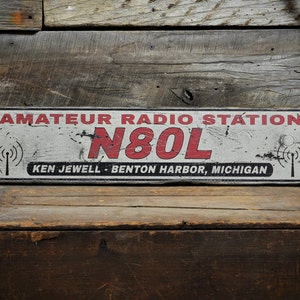 May include: A rectangular sign with a distressed white finish, featuring the red text AMATEUR RADIO STATION, the call sign N80L in large red letters, and the black text KEN JEWELL - BENTON HARBOR, MICHIGAN, flanked by two black radio tower icons.