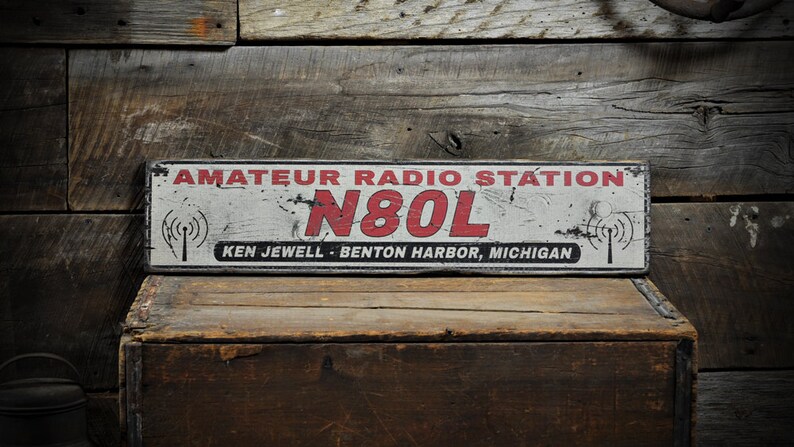 May include: A rectangular sign with a distressed white finish, featuring the red text AMATEUR RADIO STATION, the call sign N80L in large red letters, and the black text KEN JEWELL - BENTON HARBOR, MICHIGAN, flanked by two black radio tower icons.