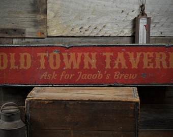 Può includere: Un cartello in legno rosso con cornice nera e lettere bianche che recita "Old Town Tavern" con le parole "Ask for Jacob's Brew" in lettere bianche più piccole sotto. Il cartello è appeso a una parete di legno rustica.