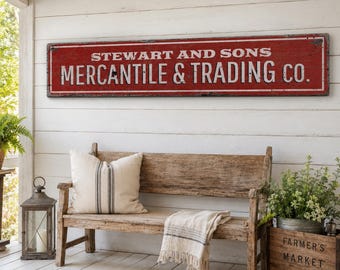 Op de afbeelding: Een lang, rechthoekig, verweerd rood houten bord met een witte rand en witte tekst die luidt STEWART AND SONS MERCANTILE & TRADING CO. Het bord heeft een verweerde afwerking met zichtbare gebruikssporen langs de randen en het oppervlak.
