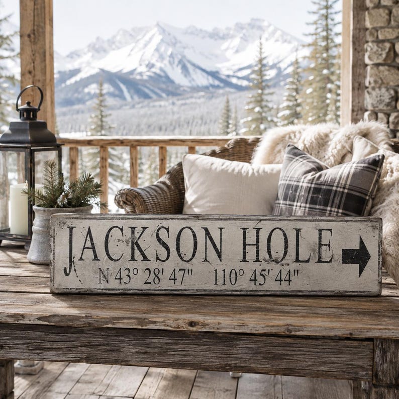 May include: A rectangular wooden sign with a distressed white finish and black lettering that reads JACKSON HOLE followed by the coordinates N 43&deg; 28' 47" 110&deg; 45' 44" and a black arrow pointing to the right side of the sign.