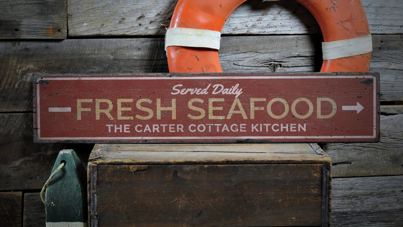 May include: A red wooden sign with white lettering that reads "Served Daily - FRESH SEAFOOD - The Carter Cottage Kitchen". The sign has a distressed look and is hanging on a wooden wall.