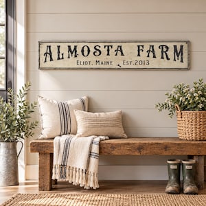 May include: A rectangular wall sign with a distressed cream finish and black lettering that reads ALMOSTA FARM, ELIOT, MAINE, EST. 2013. The sign features a thin black border and is mounted horizontally on a light-colored horizontal plank wall.