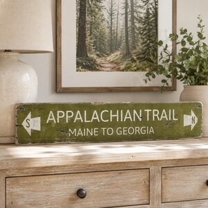 May include: A rectangular olive green wooden sign with distressed edges featuring white text that reads APPALACHIAN TRAIL and MAINE TO GEORGIA. The sign includes white directional arrows on both ends, with an S on the left and an N on the right.