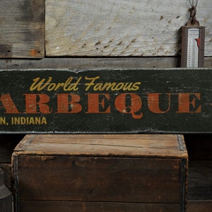 May include: A weathered wooden sign with the words "World Famous BARBEQUE" in orange lettering. The sign also includes "FRANKLIN, INDIANA" and an arrow pointing right. The sign is set against a rustic wooden background.
