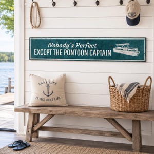 Op de afbeelding: Een rechthoekig blauwgroen wandbord met witte tekst Nobody's Perfect EXCEPT THE PONTOON CAPTAIN en een witte lijntekening van een pontonboot. Daaronder ligt een beige kussen met een zwarte anker-afbeelding en de tekst LAKE LIFE IS THE BEST LIFE.