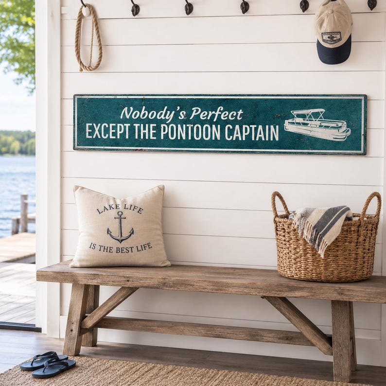 Op de afbeelding: Een rechthoekig blauwgroen wandbord met witte tekst Nobody's Perfect EXCEPT THE PONTOON CAPTAIN en een witte lijntekening van een pontonboot. Daaronder ligt een beige kussen met een zwarte anker-afbeelding en de tekst LAKE LIFE IS THE BEST LIFE.