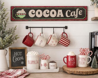 May include: A horizontal black sign with red borders reading COCOA Cafe, a black metal rack with four hanging mugs, a wooden sign reading HOT Cocoa, a white canister labelled MARSHMALLOWS, two small white containers, and two mugs labelled WARM & COZY and PEPPERMINT MOCHA.