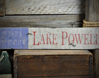 Könnte beinhalten: Ein rustikales Holzschild mit den Worten "Lake Powell" in Rot auf weißem Hintergrund. Das Schild enthält auch die Koordinaten "37° 3' 9.7" N -111° 17' 47.1" W" in Blau auf weißem Hintergrund.