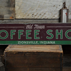 May include: A rustic wooden sign with the text "Old Town Coffee Shop" in white lettering on a maroon background. The words "Zionsville, Indiana" are written in white on a green stripe at the bottom of the sign.