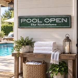 May include: A rectangular green sign with white distressed lettering that reads POOL OPEN and The Richardsons. The sign is mounted on a white horizontal paneled wall above a wooden bench holding white towels and a metal lantern.