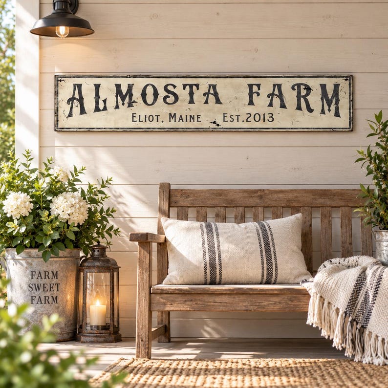 May include: A rectangular wall sign with a distressed cream finish and black lettering that reads ALMOSTA FARM, ELIOT, MAINE, EST. 2013. The sign is mounted on a horizontal wood-paneled wall above a wooden bench with a striped pillow and a throw blanket.