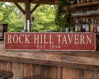 Può includere: Un'insegna rettangolare in legno con finitura rossa usurata e scritte bianche che riportano ROCK HILL TAVERN e EST. 2014. L'insegna presenta una texture invecchiata ed è posizionata orizzontalmente su un bancone da bar in legno.