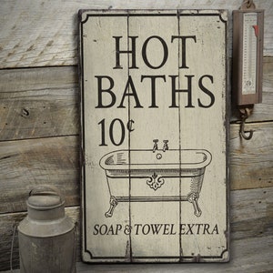 May include: A vintage-style wooden sign with white paint and black lettering advertising "HOT BATHS 10¢" with a drawing of a bathtub. The sign also says "SOAP & TOWEL EXTRA".