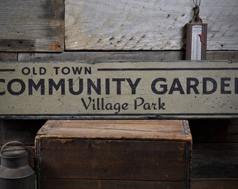 Puede incluir: Un letrero de madera desgastado con letras negras que dice "Old Town Community Garden Village Park". El letrero está colgado en una pared de madera rústica.