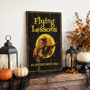 May include: A rectangular black wooden sign with vertical planks featuring the yellow text Flying Lessons and By Appointment Only. The center displays a graphic of a witch riding a broomstick in front of a large yellow moon with orange accents.