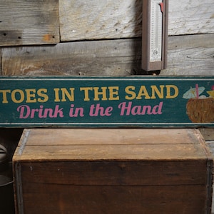 May include: A rustic wooden sign with a teal background and white lettering. The sign reads "Toes in the Sand Drink in the Hand". A colorful illustration of a coconut drink with a straw and umbrella is on the right side of the sign.