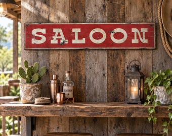 May include: A rectangular red wooden sign with the word SALOON printed in large white capital letters. The sign features a distressed, weathered finish with visible paint chips and worn edges, mounted horizontally on a rustic wood plank wall.