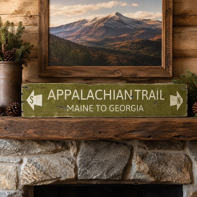 May include: A rectangular green wooden sign with distressed edges featuring the white text APPALACHIAN TRAIL and MAINE TO GEORGIA. The sign includes white directional arrows on both ends, with the letter S on the left and N on the right.