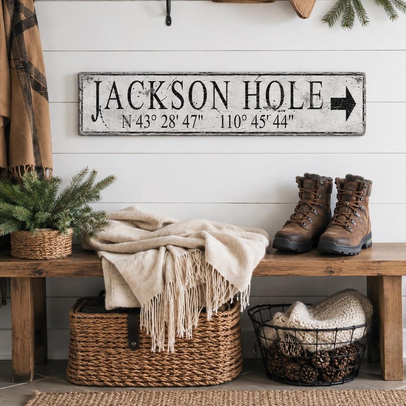 May include: A rectangular white sign with a distressed finish featuring the black text JACKSON HOLE, the coordinates N 43&deg; 28' 47" 110&deg; 45' 44", and a black arrow pointing to the right, mounted on a horizontal white paneled wall.