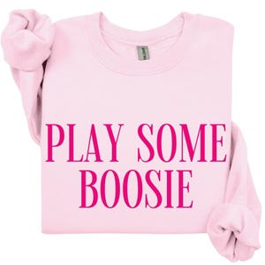 May include: A light pink sweatshirt with the phrase "PLAY SOME BOOSIE" printed in a vibrant pink font. The sweatshirt has long sleeves and a crew neck. The Gildan brand tag is visible.