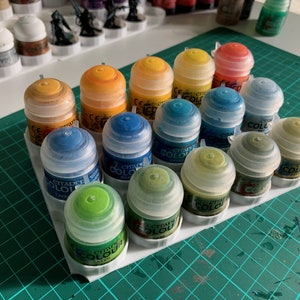 May include: A white plastic paint pot holder with 15 small paint bottles. The bottles are clear with a variety of colours including yellow, orange, green, blue, and brown. The bottles are labelled "Citadel Colour".
