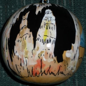 May include: A round, brown, ceramic ornament with black, white, and yellow paint depicting a building. The ornament has a gold string for hanging and the signature "Hildegard" is written on the bottom.