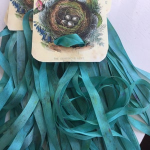 May include: A collection of teal satin ribbons attached to a rectangular card featuring a vintage botanical illustration of a bird nest with eggs and pink flowers, labeled with the text THE CHAFFINCH'S NEST, showing a smooth, lustrous texture.