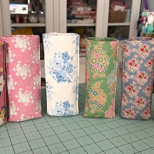 May include: Six fabric-covered cylindrical containers in various floral patterns. The colors include pink, white, blue, green, and yellow. The containers are arranged on a green cutting mat.