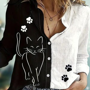 Puede incluir: Camisa abotonada con diseño dividido, con un lado negro con un contorno de gato blanco y huellas de patas, y un lado blanco con huellas de patas negras. La camisa tiene mangas largas y un corte relajado.