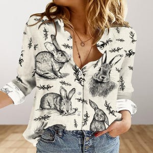 Puede incluir: Una camisa blanca de manga larga con botones y estampado de conejo blanco y negro. La camisa tiene cuello y cierre de botones en la parte delantera.