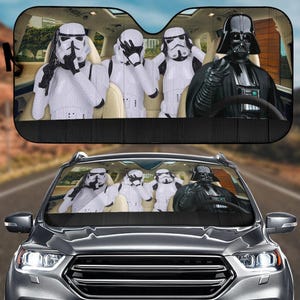 May include: Car sunshade with a Star Wars design. The image shows Darth Vader driving with three Stormtroopers in the front seats. The sunshade is black with a printed image of the characters inside a car.