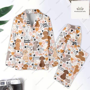 Op de afbeelding: Een witte pyjamaset met een lang mouwen shirt en broek. Het shirt heeft een knoopsluiting en een zakje op de linker borst. Het shirt en de broek hebben een herhalend patroon van bruine en witte poedels, harten, botten en de tekst "I Dog Lover", "Easily Distracted by Dogs", "Life is Better with Dogs", en "Poodles Poodles".