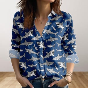 May include: A navy blue long-sleeved shirt with a repeating pattern of grey and white sharks. The shirt has a V-neck and buttoned cuffs. The shark illustrations vary in size and pose, creating a dynamic design.