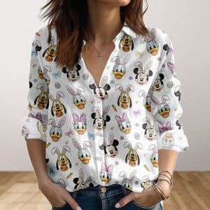 May include: White button-down shirt featuring a repeating pattern of Disney characters wearing bunny ears. Mickey Mouse, Minnie Mouse, Donald Duck, and Pluto are depicted with Easter eggs, carrots, and bows. The long-sleeved shirt has a V-neck.