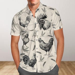 Puede incluir: Una camisa blanca de manga corta con botones y estampado de gallo en blanco y negro. La camisa tiene un corte holgado y un cuello clásico.