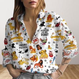 Puede incluir: Camisa blanca de botones con un estampado colorido de personajes de la película El Rey León. El estampado incluye a Simba, Nala, Mufasa, Pumbaa, Timon y otros personajes. La camisa tiene cuello en V y mangas largas. El texto "¡Estoy trabajando en mi rugido!" está impreso en la camisa.