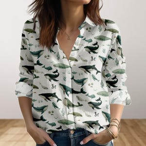Könnte beinhalten: Weißes Button-Down-Hemd mit Walmuster in Grün- und Schwarztönen. Das langärmlige Hemd hat einen klassischen Kragen und einen Knopfverschluss. Das Waldesign umfasst verschiedene Walarten und Wasserpflanzen.