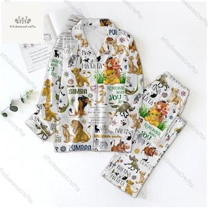 Puede incluir: Pijama blanco con estampado de El Rey León. La parte superior tiene cierre de botones y mangas largas. El pantalón tiene cintura elástica. El estampado presenta a Simba, Timón, Pumba y otros personajes de la película. El texto "Remember Who You Are" y "Hakuna Matata" también están incluidos en el estampado.