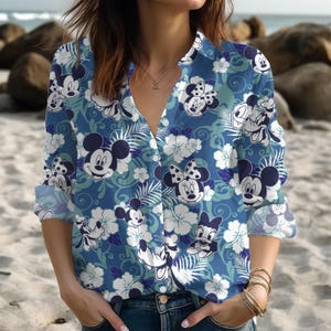 Op de afbeelding: Blauw overhemd met knopen met een patroon van Mickey en Minnie Mouse, witte bloemen en groene bladeren. Het overhemd heeft lange mouwen en een kraag. De achtergrond is lichtblauw.
