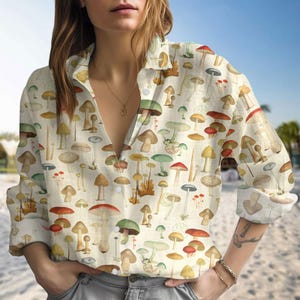 May include: A cream-coloured button-down shirt featuring a repeating pattern of various mushrooms in shades of red, green, brown, and yellow. The long-sleeved shirt has a collared neckline and is unbuttoned at the top.