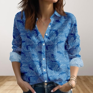 Könnte beinhalten: Blaues Button-Down-Shirt mit Hai- und Wellenmuster. Das langärmlige Hemd hat weiße Knöpfe und Manschetten. Das Design zeigt ein sich wiederholendes Muster aus Haien und wellenförmigen Linien in Weiß auf blauem Hintergrund.