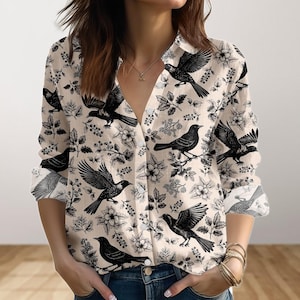 May include: A button-down shirt with a cream background and a black bird and floral pattern. The long-sleeved shirt has a collared neckline and is partially unbuttoned. The sleeves are rolled up, and the shirt is paired with blue jeans.