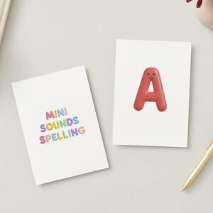 May include: Two white rectangular cards. One card features the colorful text MINI SOUNDS SPELLING. The other card displays a large, three-dimensional, glossy red letter A with a small, simple smiling face printed on its surface.