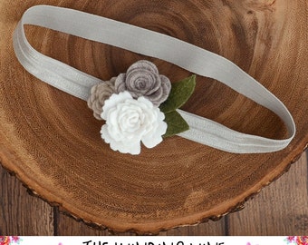 May include: A gray elastic headband with a felt flower embellishment. The flower is made of three layers of felt in shades of brown, gray, and white. The flower is accented with a green felt leaf.