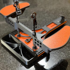 May include: A black and orange metal tool with two orange pads and two adjustable hooks. The tool has a rectangular base and a central support structure with multiple holes. The pads are textured and angled.